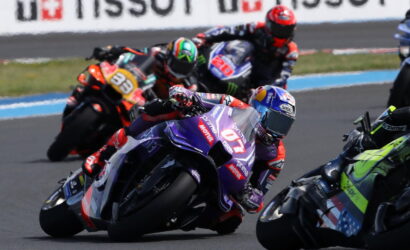 MotoGP riders lean into a corner on a race track, purple foreground bike #77 with sponsor decals.