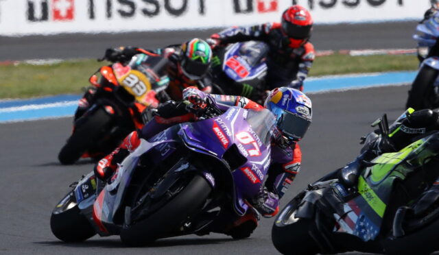 MotoGP riders lean into a corner on a race track, purple foreground bike #77 with sponsor decals.