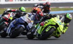 Group of motorcycle racers lean into a turn on a race track, neon-green bike at the front center.
