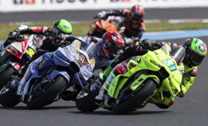 Group of motorcycle racers lean into a turn on a race track, neon-green bike at the front center.