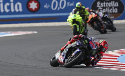Motorcycle racer in red gear leaning into a sharp turn on a race track, blue sponsor-labeled bike in foreground with a group of riders behind.
