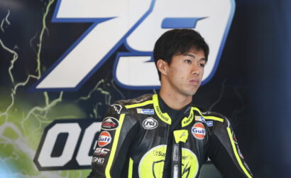 Young motorcycle racer in a black neon-yellow racing suit stands on a stage with a large blue backdrop behind him, looking to the side, sponsor patches visible on the suit.