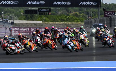Group of colorful MotoGP riders lean into a sharp turn on a racetrack, banners reading MotoGP and Qatar Airways in the background.
