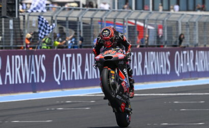 Motorcycle racer lifts front wheel on a race track, performing a wheelie with a sponsor-laden bike and Qatar Airways banners in the background.
