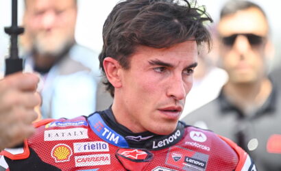 Close-up of a male motorcycle racer in a red sponsor-labeled racing suit with a focused expression.