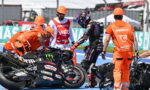 Rider in black leathers stands beside a downed race bike while crew in orange uniforms assist on a blue pit lane colorfully branded with logos nearby.