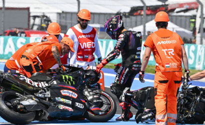 Rider in black leathers stands beside a downed race bike while crew in orange uniforms assist on a blue pit lane colorfully branded with logos nearby.