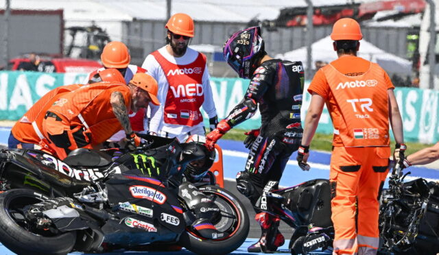 Rider in black leathers stands beside a downed race bike while crew in orange uniforms assist on a blue pit lane colorfully branded with logos nearby.