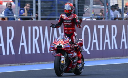 MotoGP rider in red Ducati suit on a wet track celebrating with arms raised beside a purple barrier with 'QATAR AIRWAYS' text behind him/ her on a red Ducati bike still on the track fence in a pit area.