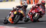 Two motorcycle racers lean into a left-hand turn on a racetrack, props and sponsor logos visible on their red and orange bikes.