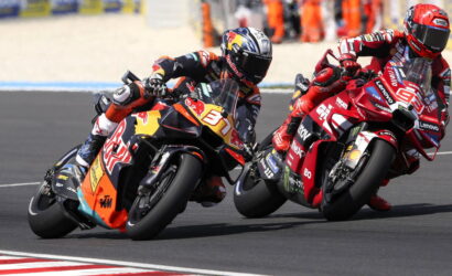Two motorcycle racers lean into a left-hand turn on a racetrack, props and sponsor logos visible on their red and orange bikes.
