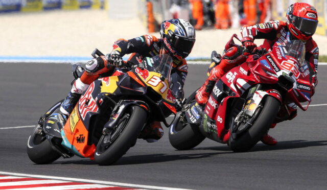 Two motorcycle racers lean into a left-hand turn on a racetrack, props and sponsor logos visible on their red and orange bikes.