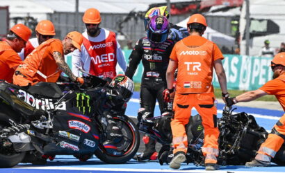 Pit crew in orange uniforms helps an injured rider in black leathers after a crash on the track, with a damaged motorcycle nearby.