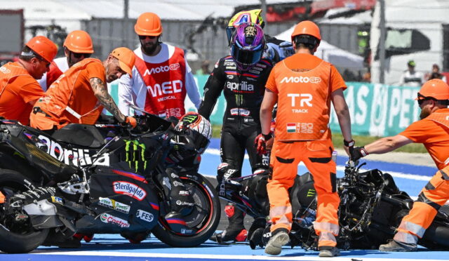Pit crew in orange uniforms helps an injured rider in black leathers after a crash on the track, with a damaged motorcycle nearby.