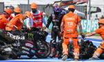 Pit crew in orange safety suits assist a fallen motorcycle on the racetrack pit lane; rider in black leathers and colorful helmet watches nearby, with MotoGP branding visible.
