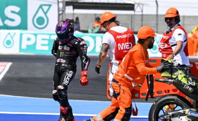 MotoGP rider in black Aprilia leathers walks away from pit crew during a race stop on the track.