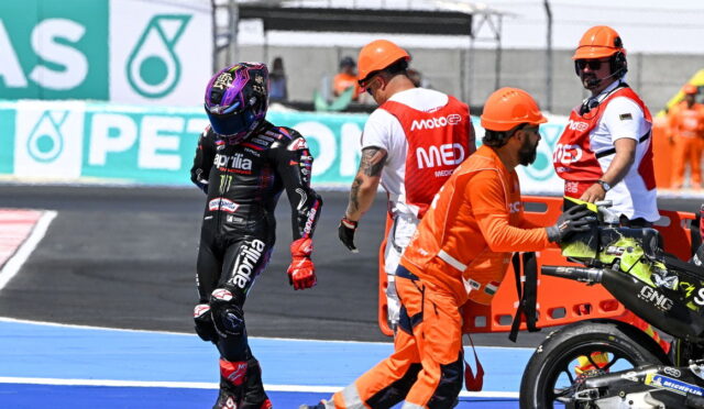 MotoGP rider in black Aprilia leathers walks away from pit crew during a race stop on the track.