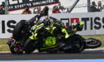 Motorcycle racer in neon-yellow suit crashes on the racetrack, leaning over another bike as fans photograph in the background.
