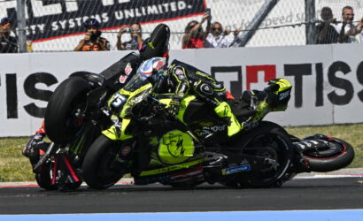 Motorcycle racer in neon-yellow suit crashes on the racetrack, leaning over another bike as fans photograph in the background.