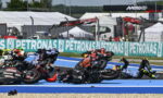 Riders in a motorcycle race crash and fall onto a blue race track, with several bikes piled on top of each other.