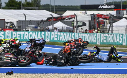Riders in a motorcycle race crash and fall onto a blue race track, with several bikes piled on top of each other.
