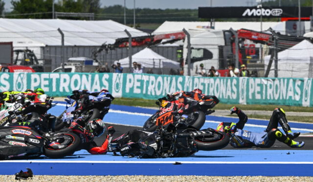 Riders in a motorcycle race crash and fall onto a blue race track, with several bikes piled on top of each other.