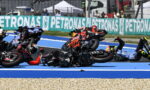 Motorcycle racers crash on a blue racetrack, several bikes toppled, riders skidding amid debris as spectators watch behind a Petronas banner.
