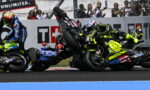 MotoGP crash: three riders in neon gear tumble on the track as bikes skid and rear wheels lift, spectators behind a fence watching.