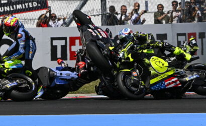 MotoGP crash: three riders in neon gear tumble on the track as bikes skid and rear wheels lift, spectators behind a fence watching.