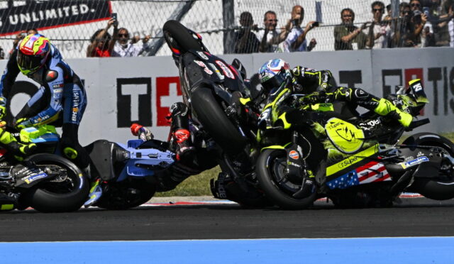 MotoGP crash: three riders in neon gear tumble on the track as bikes skid and rear wheels lift, spectators behind a fence watching.