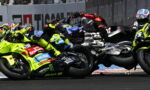 Motorcycle race in a tight pack, neon-yellow bikes and riders leaning into a corner as they collide and slide on the track momentarily.