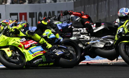 Motorcycle race in a tight pack, neon-yellow bikes and riders leaning into a corner as they collide and slide on the track momentarily.