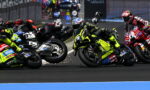 Motorcycle racers lean into a sharp corner in a crowded race, sparks flying as bikes slide wheels side-by-side across the track.