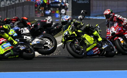 Motorcycle racers lean into a sharp corner in a crowded race, sparks flying as bikes slide wheels side-by-side across the track.