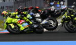 Group of motorcycle racers lean into a sharp turn on a racetrack, wearing bright yellow-green suits and helmets; sparks fly from the tires.