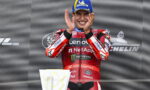 Motorsport rider on a podium clapping, wearing a red Lenovo NetApp suit and a blue Michelin cap, smiling.