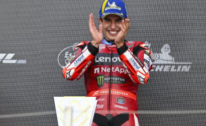 Motorsport rider on a podium clapping, wearing a red Lenovo NetApp suit and a blue Michelin cap, smiling.