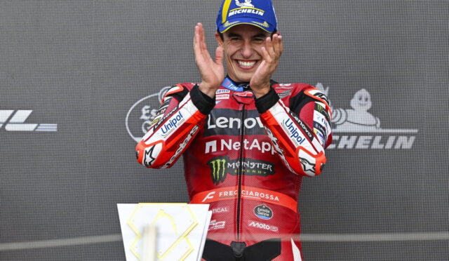 Motorsport rider on a podium clapping, wearing a red Lenovo NetApp suit and a blue Michelin cap, smiling.