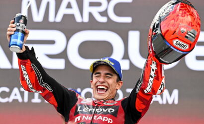 MotoGP rider in a red racing suit celebrates on a podium, raising a beer can in one hand and his helmet in the other, smiling broadly.