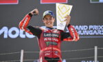 MotoGP rider in a red Lenovo NetApp racing suit on the podium, raising a trophy with one hand and fist, smiling.