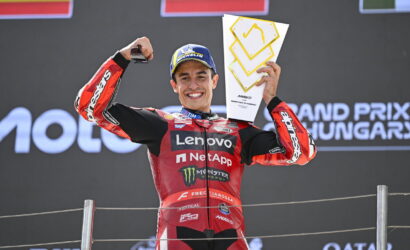 MotoGP rider in a red Lenovo NetApp racing suit on the podium, raising a trophy with one hand and fist, smiling.