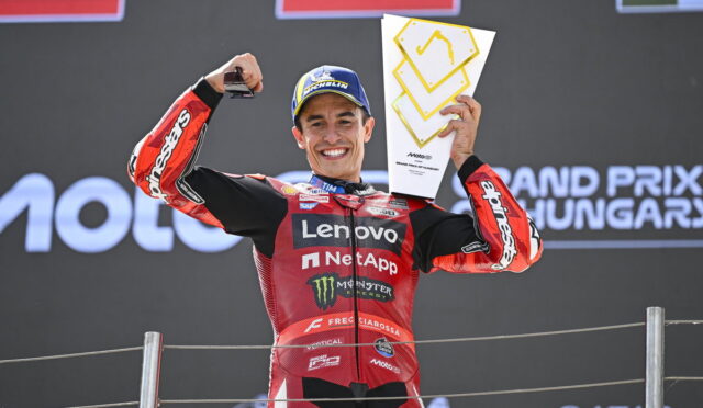 MotoGP rider in a red Lenovo NetApp racing suit on the podium, raising a trophy with one hand and fist, smiling.