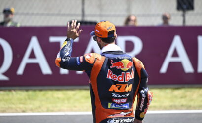 Motorcycle racer in a Red Bull KTM suit waves to the crowd from the track, back view with sponsor logos visible.