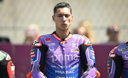 MotoGP rider in purple Pramac Yamaha leathers poses on track with arms crossed and a focused expression.