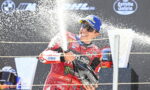 Motorcycle racer in a red suit sprays champagne on the podium, wearing a blue cap and mirrored sunglasses, celebrating victory.