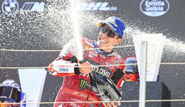 Motorcycle racer in a red suit sprays champagne on the podium, wearing a blue cap and mirrored sunglasses, celebrating victory.