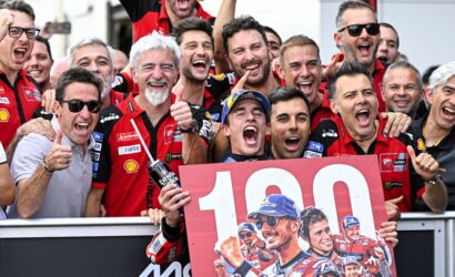 Group of cheering motorbike racers wearing red team shirts celebrate a win, holding a large 100 banner with a bike rider image.