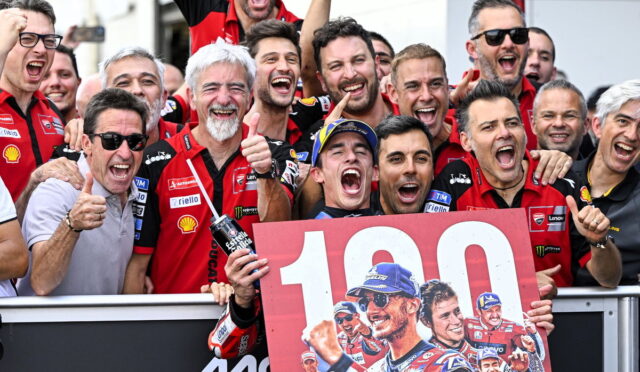 Group of cheering motorbike racers wearing red team shirts celebrate a win, holding a large 100 banner with a bike rider image.