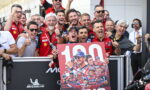 Group of race team members celebrating behind a podium-style banner that reads '100 WINS' outdoors. Thousands of people celebrate victory vibes conveyed by smiles and thumbs up.