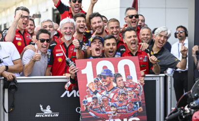 Group of race team members celebrating behind a podium-style banner that reads '100 WINS' outdoors. Thousands of people celebrate victory vibes conveyed by smiles and thumbs up.